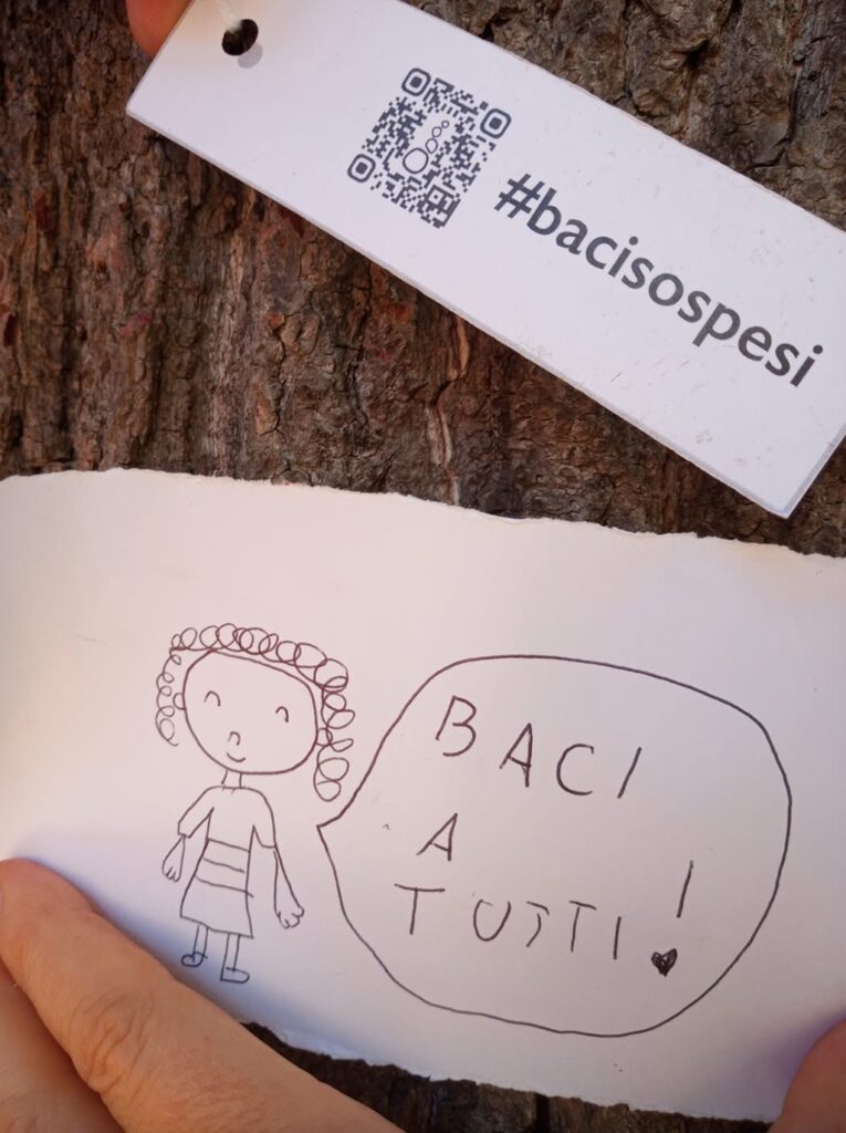 A hand holding a drawn message beside red Baci Sospesi forms on a tree.
