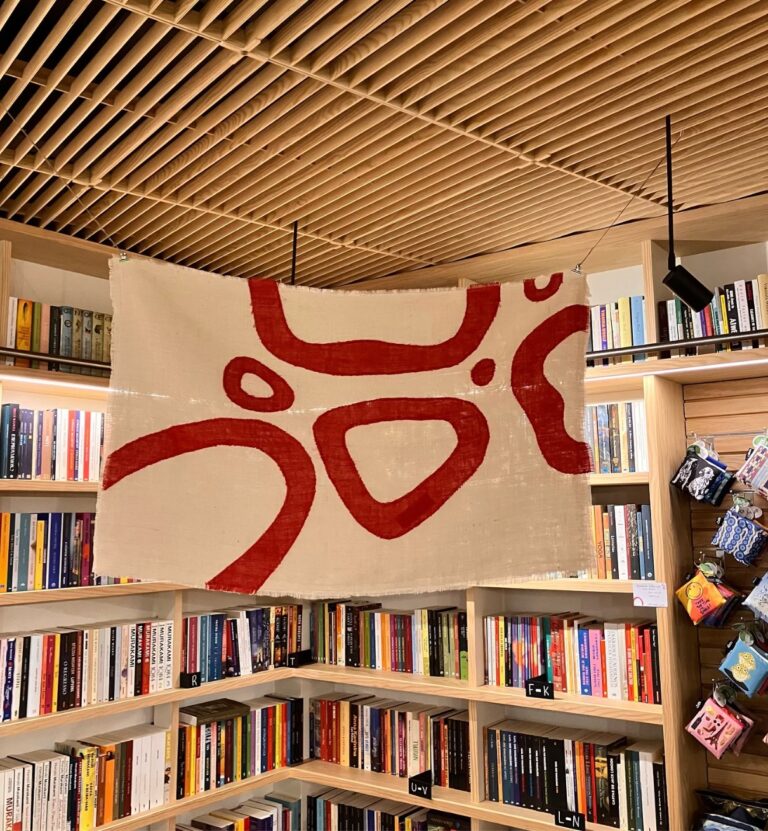 The complete handmade Baci Sospesi flag prototype in jute, displayed among books at Livraria Arquivo in Leiria.